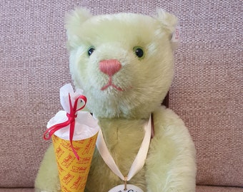 Könnte beinhalten: Ein grüner Steiff Club Edition Teddybär mit rosa Nase und rosa Pfoten. Der Bär hält einen gelben und roten Papierkegel mit einem roten Band. Der Bär trägt ein weißes Band mit einem runden weißen Etikett, auf dem "Steiff Club Edition 1998" steht.