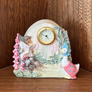 May include: A decorative clock with a charming scene of a mouse reading and a duck in a bonnet. The clock face is gold-coloured with black numerals. The scene includes pink flowers and is set in a garden. The clock is a decorative item.