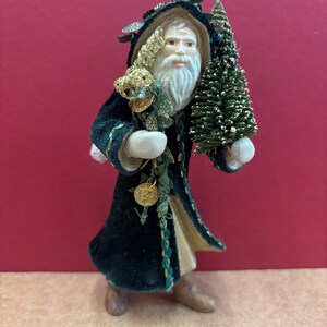 May include: A Santa Claus figurine in a dark green velvet coat with gold trim and hood, holding a small Christmas tree and a gold ornament. The figure has a long white beard and stands on a brown base. The figurine is a festive Christmas decoration.