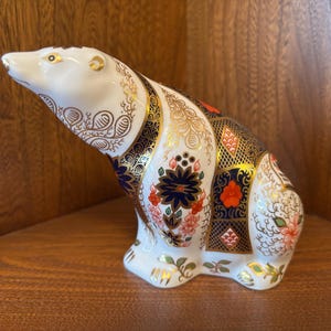 Puede incluir: Una figurita de oso polar de porcelana blanca con un diseño estampado en azul, rojo y dorado. El oso está sentado en sus ancas con la cabeza inclinada hacia arriba.