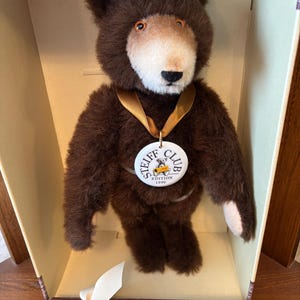 Peut inclure: Un ours en peluche marron avec un visage brun clair et un ruban doré autour du cou. L'ours porte un médaillon Steiff Club Edition 1996. L'ours est dans une boîte avec une étiquette indiquant "Made in Germany by Steiff in Ohr".