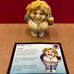 May include: A figurine of a woman holding a beer mug. The figurine is made of a light brown material and has a cartoonish style. The text on the card reads "Beer Babe (the waitress)" and "Fun Facts".