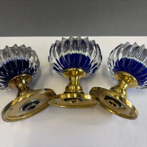 Może przedstawiać: Trzy zabytkowe gałki do drzwi z przezroczystymi, żebrowanymi szklanymi wierzchami i głębokim niebieskim wnętrzem. Gałki są osadzone na mosiężnych podstawach ze złotym wykończeniem. Układ znajduje się na białej powierzchni.