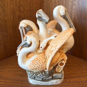 May include: A ceramic sculpture of multiple pelicans huddled together. The birds are primarily cream-colored with tan accents on their wings and beaks. The base of the sculpture has the text "BIRD IS THE". The sculpture is displayed on a wooden surface.