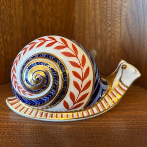 Puede incluir: Figura decorativa de caracol de cerámica. La concha es blanca con detalles de hojas rojas y un diseño en espiral azul y dorado. La base y la cabeza son blancas con detalles dorados. Sobre una superficie de madera.