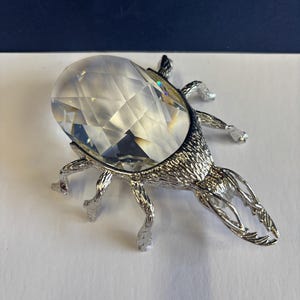 Könnte beinhalten: Eine dekorative silberfarbene Käferfigur mit einem großen, facettierten, klaren Kristallkörper. Der Käfer hat detaillierte Beine, Fühler und Mandibeln. Der Kristall reflektiert das Licht. Die Figur steht auf einer weißen Oberfläche.