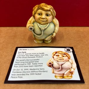 May include: A figurine of a female doctor wearing a white coat and a stethoscope. The figurine is holding a small book. The figurine is made of a light brown material. The figurine is standing on a white background. The text on the card reads: "Life Saver (the doctor) Fun Facts The U.S. spends more on health care than any other nation, over 14% of the Gross Domestic Product. The world's first successful heart-lung transplant was performed in 1981. Today more than 2510 have been reported. On Oct. 15, 1999, Medecins Sans Frontieres (Doctors Without Borders) was awarded the 1999 Nobel Peace Prize."