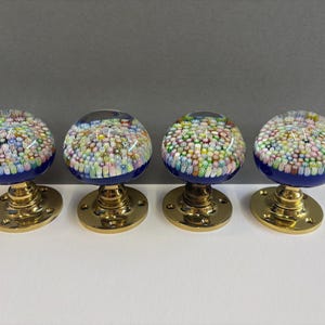 Op de afbeelding: Vier decoratieve deurknoppen met een kleurrijk, mozaïekachtig ontwerp. Elke knop heeft een heldere, afgeronde bovenkant gevuld met kleine, veelkleurige stukjes, geplaatst op een donkerblauwe basis en een goudkleurige metalen basis. De knoppen zijn in een rij gerangschikt.