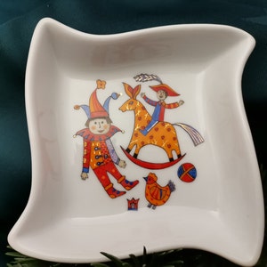 Könnte beinhalten: Weiße Keramik-Schale mit einem fantasievollen Design, das einen Narren, ein Kind auf einem Schaukelpferd und ein Huhn zeigt. Das Design ist in leuchtenden Farben gehalten, darunter Rot, Orange, Gelb, Blau und Grün.