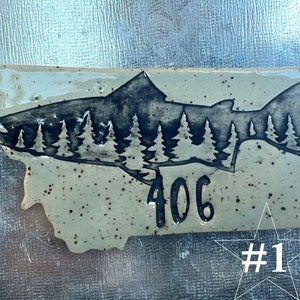 Puede incluir: Un azulejo de cerámica con forma del estado de Montana con una silueta negra de un salmón con un bosque de árboles dentro de la silueta. El número 406 está impreso debajo del salmón. El azulejo tiene un fondo beige moteado. El azulejo está etiquetado como #1.