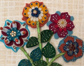 May include: A vibrant felt artwork depicting a vase of flowers. The purple vase has embroidered patterns, and the flowers are in red, orange, and blue, with green stems and leaves. The artwork is on a textured background.
