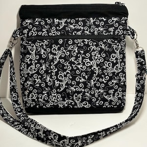 May include: Black crossbody bag with a white floral pattern. The bag has two zippered compartments and a long adjustable strap.