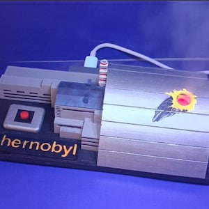 Peut inclure: Maquette de la centrale nucléaire de Tchernobyl, avec une base rectangulaire portant l'inscription "Chernobyl" en lettres dorées. Le modèle comprend un bouton, des bâtiments et une structure métallique avec un motif peint.