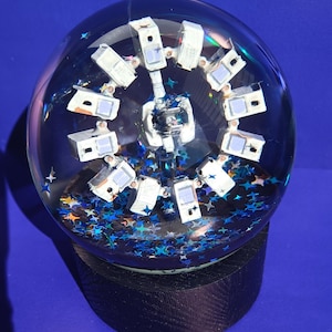 Peut inclure: Un globe de neige en verre transparent avec un modèle de vaisseau spatial blanc entouré de paillettes en forme d'étoiles bleues et argentées. Le globe repose sur une base en bois noir, sur fond bleu.