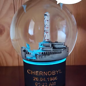 May include: A snow globe with a miniature model of the Chernobyl nuclear power plant inside. The base of the globe is black with the words "CHERNOBYL 26.04.1986 01:23 AM" inscribed in gold lettering. The globe has a blue light around the base.