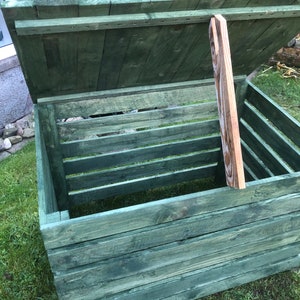 May include: A green wooden compost bin with a hinged lid. The lid is open and propped up by a wooden support.