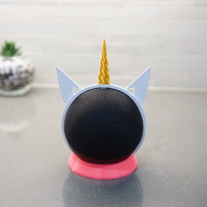 Peut inclure: Un support de haut-parleur en forme de licorne blanche avec une corne dorée et une base rose. Le haut-parleur est noir et rond.