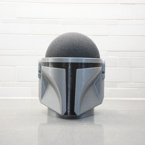 Peut inclure: Un casque gris et noir imprimé en 3D ressemblant au casque du Mandalorien de la franchise Star Wars. Le casque est posé sur une surface grise.