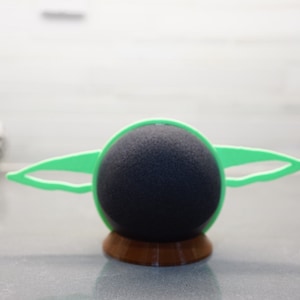 May include: A green and black 3D printed stand for a speaker, shaped like the character Baby Yoda from the Star Wars series. The stand has a round base and two ear-like protrusions.