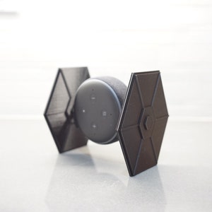 May include: A black Amazon Echo Dot smart speaker mounted on a 3D printed stand shaped like a TIE fighter from Star Wars.