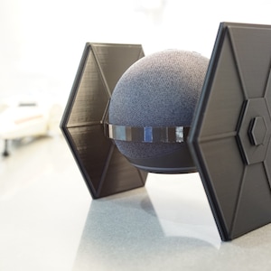 May include: A black 3D printed TIE fighter stand for a smart speaker. The stand is designed to hold a round speaker, and the wings of the TIE fighter are angled upwards.