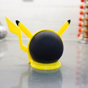 May include: A yellow Pikachu-themed speaker stand with black accents. The stand is designed to hold a round, black smart speaker.