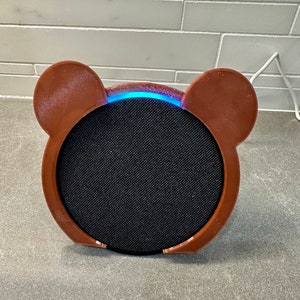 May include: A brown speaker with two ear-like protrusions on the sides. The speaker has a black fabric grille and a blue light ring around the top.