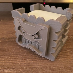 May include: A gray 3D printed note holder shaped like a monster's face with a textured surface. The holder has a square opening for holding sticky notes.