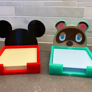 May include: Two 3D printed note holders, one with Mickey Mouse ears and a red base, the other with a tan raccoon face and a teal base.