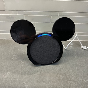 May include: Black smart speaker with Mickey Mouse ears. The speaker has a blue light ring around the top.