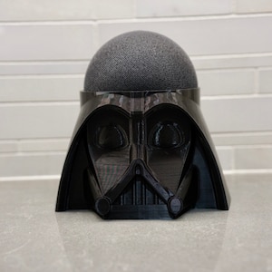 May include: A black Darth Vader-themed stand for a smart speaker. The stand is made of plastic and has a detailed design of Darth Vader's helmet.