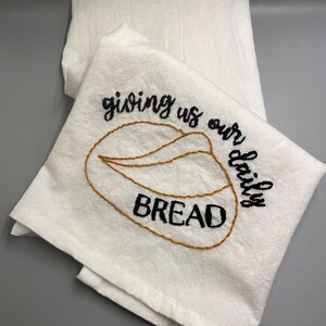 May include: White kitchen towel with embroidered text "giving us our daily bread" and a stylized image of a loaf of bread.