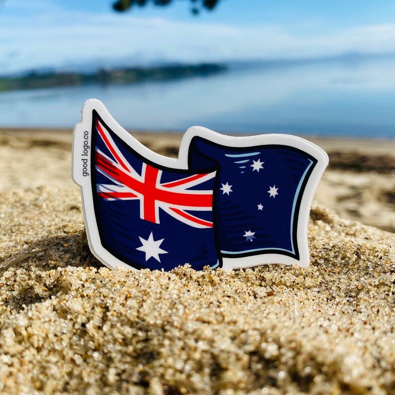 May include: A sticker of the Australian flag waving in the wind. The flag is blue, red, and white with a white star in the center and a constellation of stars in the upper right corner. The sticker is on a sandy beach with the ocean in the background. The text "good logo.co" is printed on the sticker.