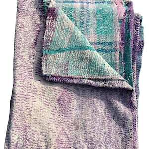 May include: A folded, multi-colored textile with a patchwork design. The top layer features a teal and purple plaid pattern, while the folded section reveals a white and purple stitched design. The edges are frayed, giving it a vintage look.