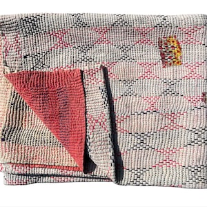 May include: Folded, handmade quilt with a geometric pattern. The quilt features a cream-colored background with a diamond pattern in red and black stitching. The reverse side is a solid coral color. A small patch of yellow and orange stitching is visible.