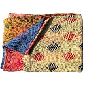 May include: A folded, multi-colored quilt with a cream-colored top featuring red and black diamond patterns. The quilt's edges reveal a blue and pink underside, and a yellow and green patterned section.