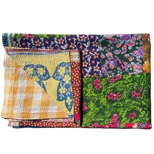 Op de afbeelding: Gevouwen patchworkquilt met een verscheidenheid aan bloemen- en geometrische patronen. De quilt heeft secties van gele ruitjes, blauwe bloemen en groene bloemenontwerpen. De randen zijn afgewerkt met een donkerblauw bloemenpatroon en rode stippen.