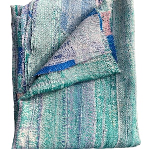 May include: A folded, multi-colored blanket with a textured, stitched appearance. The blanket features vertical stripes in shades of teal, blue, and white, with a folded section revealing a blue border and pink accents. The blanket is made of a soft material.