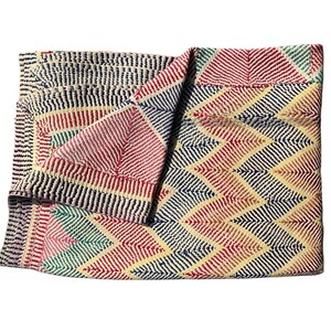 Könnte beinhalten: Gefalteter Stoff mit geometrischem Muster. Das Design zeigt ein Zickzackmuster in Rot, Marineblau, Gelb und Grün. Die Kanten haben ein Streifenmuster in Marineblau, Rot und Gelb.