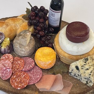 May include: A wooden serving board with a variety of cheeses, meats, and bread. There is a bottle of red wine, a bunch of grapes, and a small loaf of bread. The board is decorated with a few sprigs of greenery.