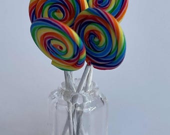 May include: A small glass jar holds a bouquet of colorful, swirled lollipops. The lollipops feature a rainbow of colors, including red, orange, yellow, green, blue, and purple. The lollipops are on white sticks.