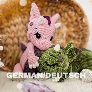 Könnte beinhalten: Zwei gehäkelte Dinosaurier-Spielzeuge. Eines ist rosa mit weißen Hörnern und einem lila Kamm, das andere ist grün mit weißen Hörnern. Die Spielzeuge sind handgefertigt und haben große schwarze Augen. Der Text "GERMAN/DEUTSCH" steht unten.