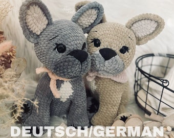 May include: Two crocheted plush French Bulldog toys, one gray and one beige, sitting on a white fuzzy surface. The toys have black noses and large black eyes.