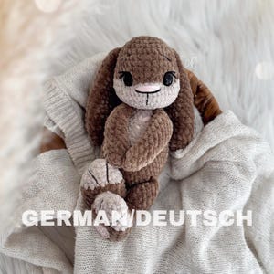 Könnte beinhalten: Ein handgehäkelter Hase in Braun- und Beigetönen. Das Stofftier hat lange Ohren, ein süßes Gesicht mit schwarzen Augen und eine rosa Nase. Der Hase liegt auf einer weichen, weißen Oberfläche.