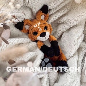 Könnte beinhalten: Ein handgefertigtes Fuchs-Plüschtier in Orange, Schwarz und Weiß, das auf einer weichen, cremefarbenen Decke liegt. Der Fuchs hat große, dunkle Augen und einen lieben Ausdruck. Der Text "GERMAN/DEUTSCH" ist sichtbar.