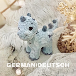 Könnte beinhalten: Ein hellblaues gehäkeltes Dinosaurierspielzeug mit schwarzen Augen und einem lächelnden Gesicht. Das Spielzeug sitzt auf einer weißen, flauschigen Oberfläche.