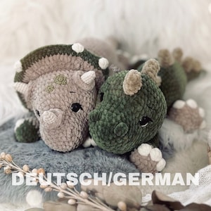 May include: Two crocheted stuffed animals, a green dinosaur and a gray triceratops, are lying on a gray fuzzy surface. The dinosaur has a beige horn and white spots. The triceratops has green spots and white spots. The text "DEUTSCH/GERMAN" is printed on the surface.