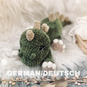 Könnte beinhalten: Ein handgefertigtes grünes und beiges Häkeldinosaurierspielzeug. Der Dinosaurier hat große schwarze Augen, beige Hörner und beige und weiße Füße. Der Text "GERMAN/DEUTSCH" befindet sich am unteren Rand des Bildes.