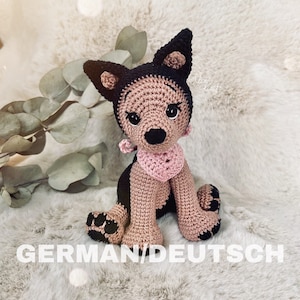 Può includere: Cane di peluche lavorato a uncinetto in marrone e nero con un collare rosa a forma di cuore. Il cane è seduto su una superficie bianca e soffice.