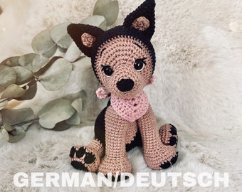 May include: Crocheted plush toy dog in brown and black with a pink heart-shaped collar. The dog is sitting on a white fluffy surface.